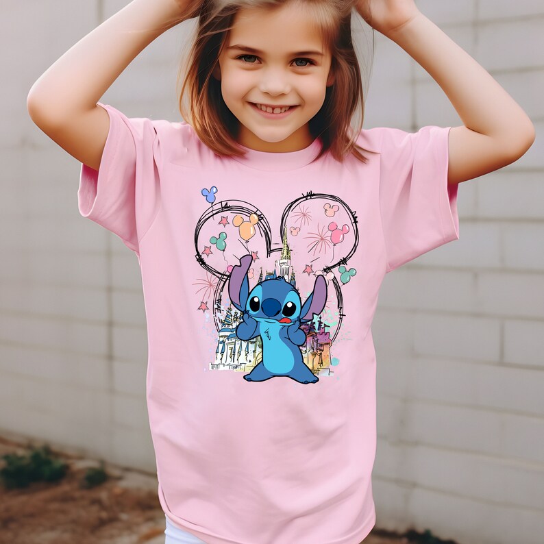 May include: A pink t-shirt with a blue Stitch character wearing Mickey Mouse ears with colorful balloons and fireworks. The shirt features a sketch of Cinderella's castle in the background.