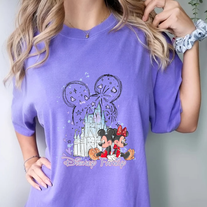 May include: Purple t-shirt featuring a graphic of Mickey Mouse and Minnie Mouse in front of Cinderella's Castle. The graphic also includes fireworks and the text "Disney Family".