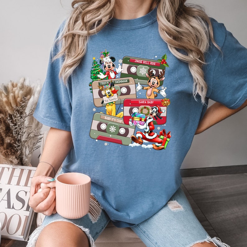 Puede incluir: Una camiseta azul con un dise&ntilde;o navide&ntilde;o vintage con Mickey Mouse y otros personajes de Disney en cintas de casete. El texto en las cintas incluye "Jingle Bell Rock", "First Snowman", "Santa Baby" y "Baby It's Cold Outside".