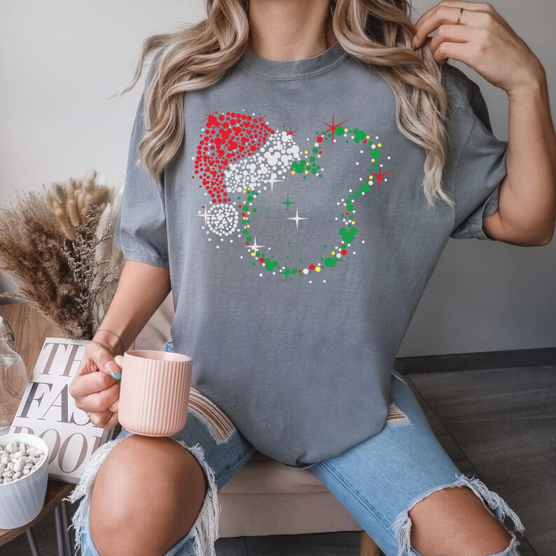 Puede incluir: Camiseta gris con un dise&ntilde;o festivo de una cabeza de rat&oacute;n con un gorro de Pap&aacute; Noel, creado con puntos rojos y blancos. La cabeza del rat&oacute;n est&aacute; rodeada por una corona verde con detalles rojos y amarillos. La camiseta la lleva una persona.