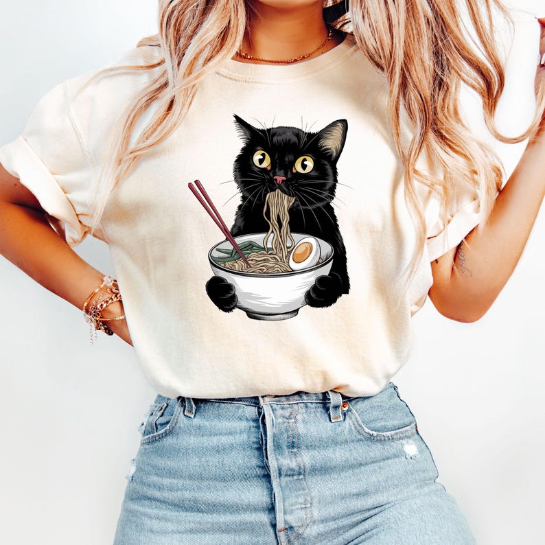 Puede incluir: Una camiseta blanca con un gato negro comiendo fideos ramen de un taz&oacute;n blanco. El gato tiene ojos verdes y sostiene palillos en sus patas.