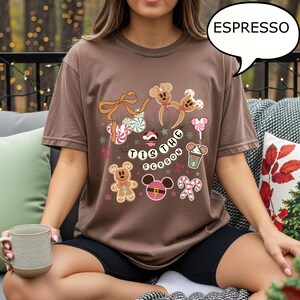 May include: Espresso-colored t-shirt featuring a festive design with gingerbread men, candy canes, and the phrase "Tis the Season." The graphic includes Mickey Mouse-inspired elements and holiday-themed illustrations. The shirt is a comfortable, casual style.
