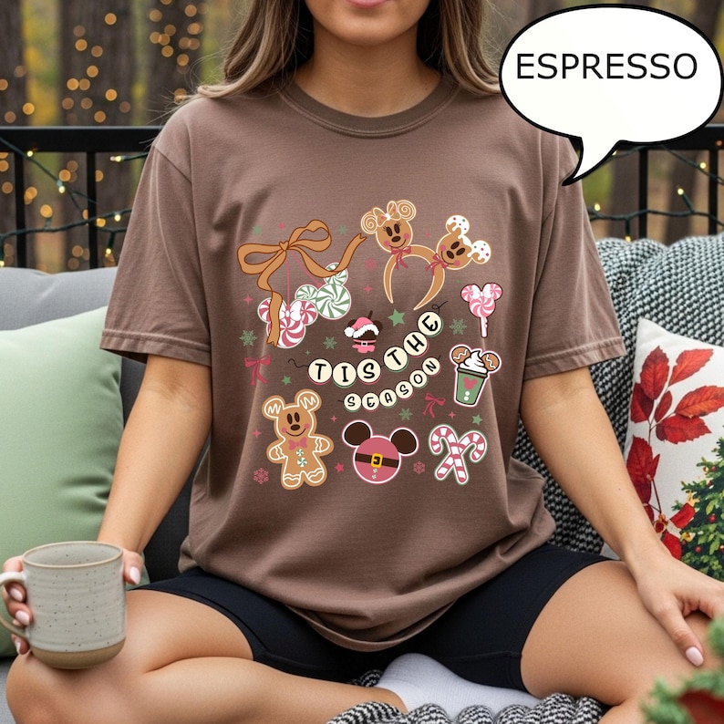 May include: Espresso-colored t-shirt featuring a festive design with gingerbread men, candy canes, and the phrase "Tis the Season." The graphic includes Mickey Mouse-inspired elements and holiday-themed illustrations. The shirt is a comfortable, casual style.