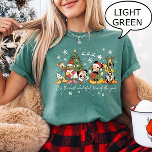 May include: Light green t-shirt featuring a Christmas design with Disney characters, a decorated tree, snowflakes, and the text "It's the most wonderful time of the year." The shirt has a relaxed fit.
