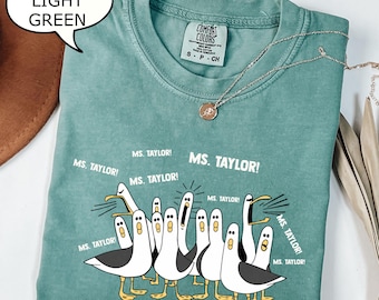 May include: Light green Comfort Colors t-shirt with a graphic of cartoon seagulls and the text "MS. TAYLOR!". The shirt has a crew neck and short sleeves. A silver necklace with a pendant is on the shirt.
