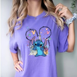 May include: Purple t-shirt with a blue Stitch character holding balloons in front of a cartoon castle. The design is inside a Mickey Mouse ear outline.