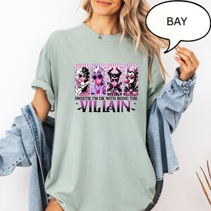 May include: Sage green t-shirt featuring a graphic of four cartoon villains with the text "Sweetie I'm OK with Being the Villain". The graphic includes characters in black, white, purple, and red. The shirt has short sleeves.