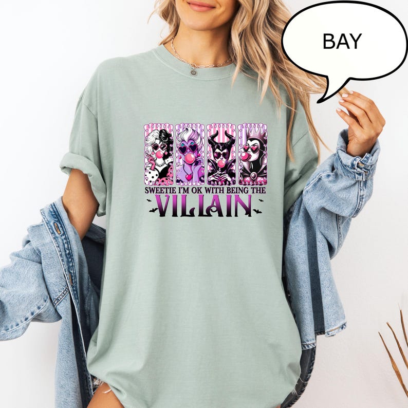 May include: Sage green t-shirt featuring a graphic of four cartoon villains with the text "Sweetie I'm OK with Being the Villain". The graphic includes characters in black, white, purple, and red. The shirt has short sleeves.