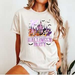 May include: A light beige t-shirt with a purple and white graphic of Mickey Mouse and friends in front of the Cinderella Castle. The text "Mickey's Not So Scary Halloween Party" is printed in purple on the shirt.