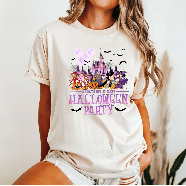 May include: A light beige t-shirt with a purple and white graphic of Mickey Mouse and friends in front of the Cinderella Castle. The text "Mickey's Not So Scary Halloween Party" is printed in purple on the shirt.