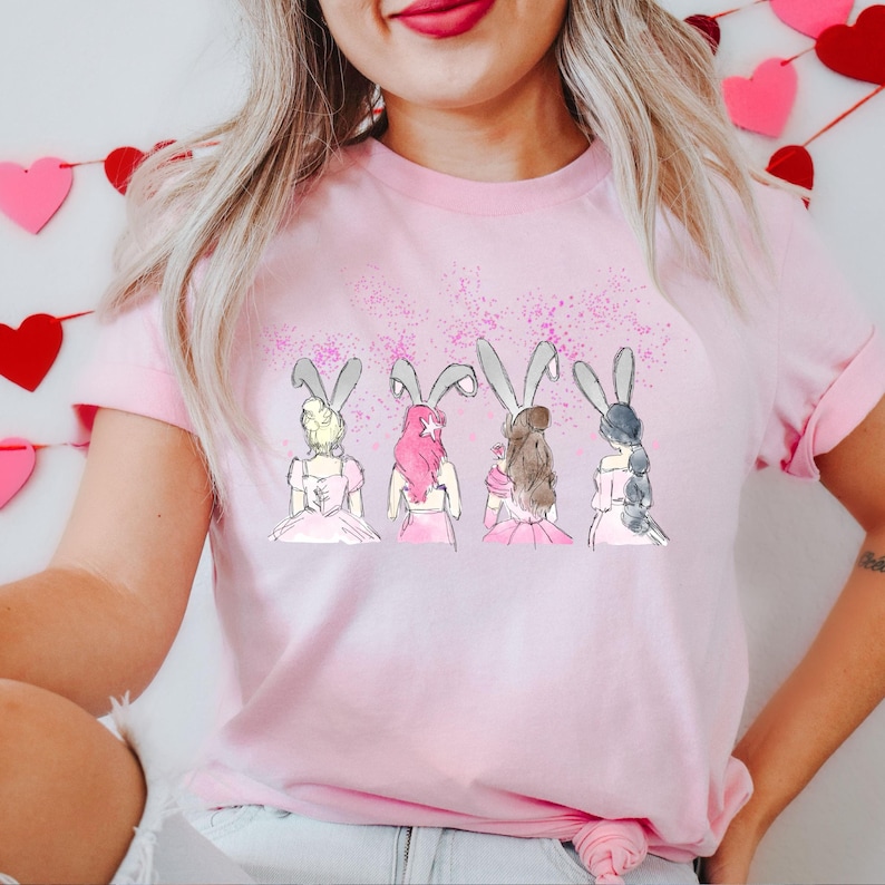 Puede incluir: Camiseta rosa con un gr&aacute;fico de cuatro mujeres con orejas de conejo y vestidos rosas. El gr&aacute;fico est&aacute; rodeado de purpurina rosa.