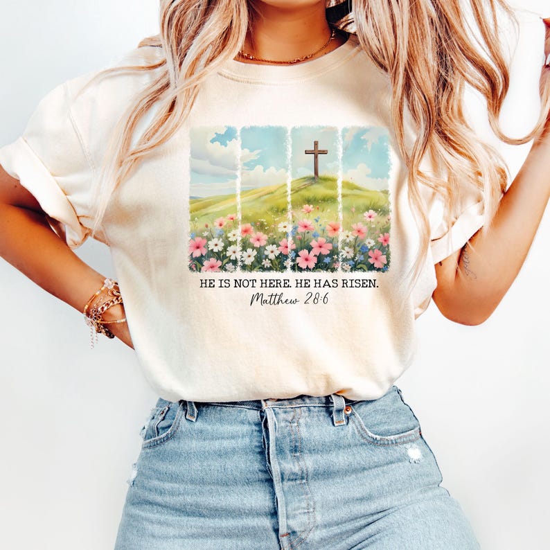 May include: White t-shirt featuring a graphic of a wooden cross on a hill with flowers. The text "He is not here. He has risen. Matthew 28:6" is printed below the graphic.