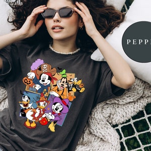 K&ouml;nnte beinhalten: Ein schwarzes T-Shirt mit einem bunten Motiv von Mickey Mouse und seinen Freunden in Halloween-Kost&uuml;men. Das Motiv zeigt Mickey Mouse als Pirat, Goofy als Geist, Donald Duck als Hexe und Pluto als Skelett. Der Text "PEPPER" ist im Hintergrund sichtbar.