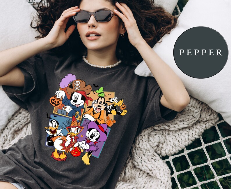 K&ouml;nnte beinhalten: Ein schwarzes T-Shirt mit einem bunten Motiv von Mickey Mouse und seinen Freunden in Halloween-Kost&uuml;men. Das Motiv zeigt Mickey Mouse als Pirat, Goofy als Geist, Donald Duck als Hexe und Pluto als Skelett. Der Text "PEPPER" ist im Hintergrund sichtbar.