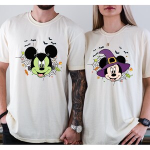 Może przedstawiać: Dwie kremowe koszulki z motywami Myszki Miki i Myszki Minnie w stylu Halloween. Jedna koszulka przedstawia Myszkę Miki jako Frankensteina, druga Myszkę Minnie w kapeluszu czarownicy. Obie koszulki mają pajęczyny, nietoperze i cukierki.