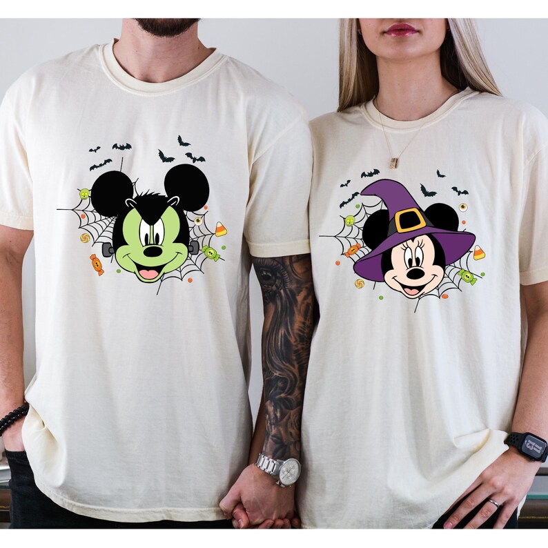 Może przedstawiać: Dwie kremowe koszulki z motywami Myszki Miki i Myszki Minnie w stylu Halloween. Jedna koszulka przedstawia Myszkę Miki jako Frankensteina, druga Myszkę Minnie w kapeluszu czarownicy. Obie koszulki mają pajęczyny, nietoperze i cukierki.