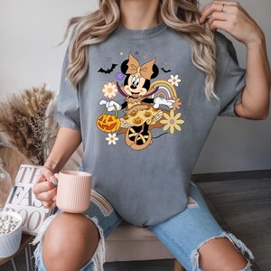 Puede incluir: Camiseta gris con un dise&ntilde;o de Minnie Mouse de Halloween. Minnie est&aacute; disfrazada y sostiene una linterna de calabaza. El dise&ntilde;o incluye flores, murci&eacute;lagos y un arco&iacute;ris. La camiseta la lleva una persona.