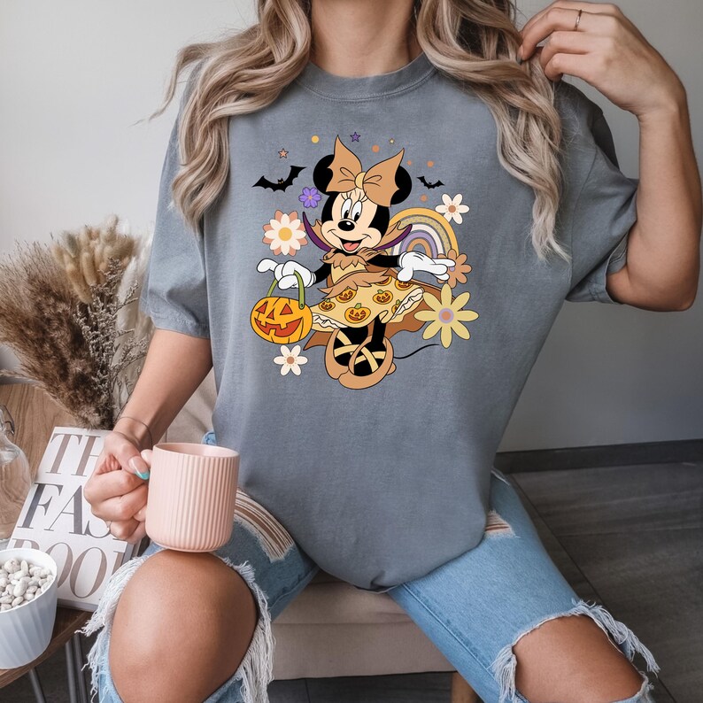 Puede incluir: Camiseta gris con un dise&ntilde;o de Minnie Mouse de Halloween. Minnie est&aacute; disfrazada y sostiene una linterna de calabaza. El dise&ntilde;o incluye flores, murci&eacute;lagos y un arco&iacute;ris. La camiseta la lleva una persona.