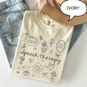 May include: Ivory-colored t-shirt with the words "Speech therapy" and various green illustrations. The shirt also features the phrases "Communication is Connection" and "Your Words Matter". The shirt is displayed with blue jeans and a tan hat.