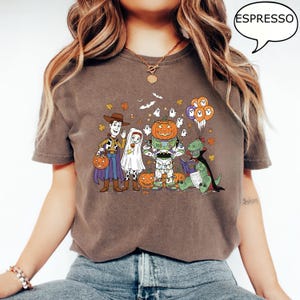 Puede incluir: Camiseta color espresso con un gr&aacute;fico de dibujos animados con tem&aacute;tica de Halloween. El dise&ntilde;o incluye personajes disfrazados de Woody, un fantasma, Buzz Lightyear con cabeza de calabaza y un dinosaurio leyendo un libro, con calabazas y hojas de oto&ntilde;o.