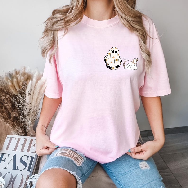 May include: A pink t-shirt featuring a graphic of a ghost wearing a polka dot sheet and walking a dog wearing a sheet. The dog is looking back at the ghost.