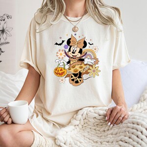Puede incluir: Camiseta de color crema con un dise&ntilde;o de Minnie Mouse con tem&aacute;tica de Halloween. El dise&ntilde;o incluye una calabaza, murci&eacute;lagos, flores y un arco&iacute;ris. Minnie Mouse est&aacute; vestida con un disfraz de Halloween. La camiseta tiene mangas cortas y un corte holgado.