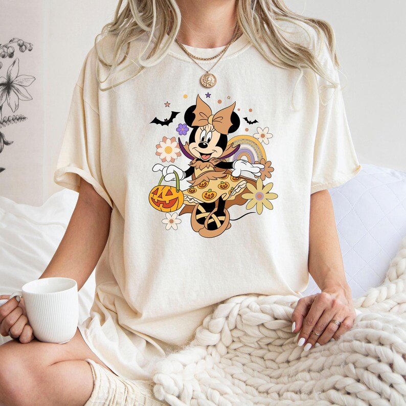 Puede incluir: Camiseta de color crema con un dise&ntilde;o de Minnie Mouse con tem&aacute;tica de Halloween. El dise&ntilde;o incluye una calabaza, murci&eacute;lagos, flores y un arco&iacute;ris. Minnie Mouse est&aacute; vestida con un disfraz de Halloween. La camiseta tiene mangas cortas y un corte holgado.