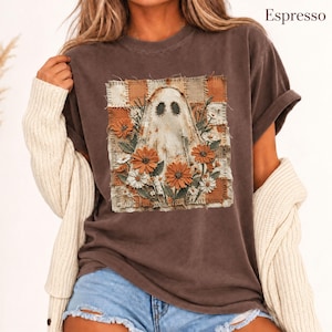 以下が含まれることがあります： エスプレッソカラーの半袖Tシャツ。中央にはパッチワーク風の背景にオレンジと白の花々に囲まれたゴーストのグラフィックが描かれています。デザインにはステッチのディテールが含まれ、右上にEspressoの文字があります。