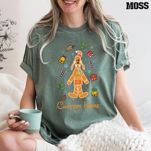 May include: A green t-shirt with a gingerbread man design. The gingerbread man is wearing a red bow tie and has a red candy cane in his hand. The design also includes candy canes, bells, and other Christmas decorations. The text "Custom Name" is printed below the design.