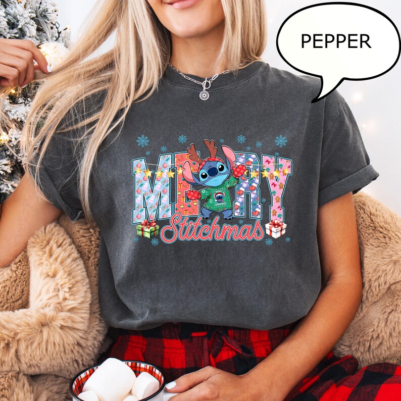 Op de afbeelding: Donkergrijs T-shirt met het woord "MERRY" in een feestelijk patroon en een stripfiguur met een kersttrui. De tekst "Stitchmas" staat onder het woord "MERRY". Het shirt heeft een relaxte pasvorm.