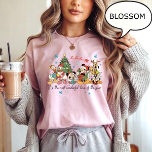 May include: A light pink long-sleeve shirt featuring a festive graphic of Disney characters, a Christmas tree, and snowflakes. The text "It's the most wonderful time of the year" is printed below the graphic.