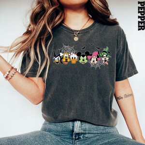 May include: A dark grey t-shirt featuring a graphic print of Mickey Mouse and his friends dressed in Halloween costumes. The design includes Mickey Mouse, Minnie Mouse, Donald Duck, Goofy, and Pluto. The graphic is surrounded by a spider web design.