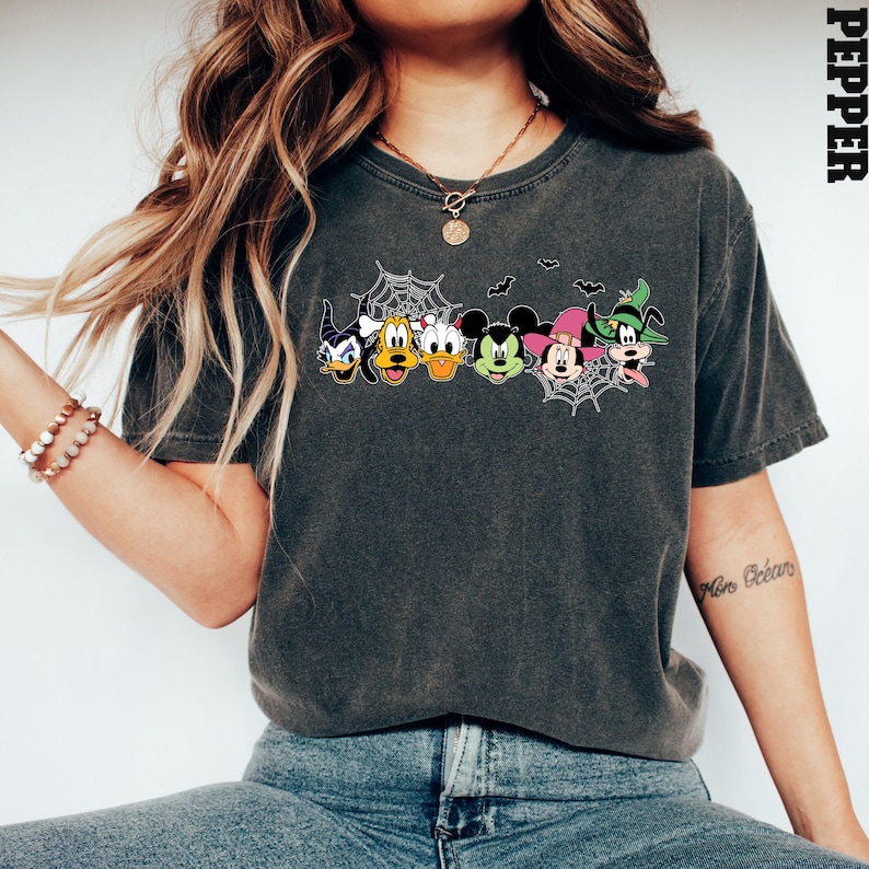 May include: A dark grey t-shirt featuring a graphic print of Mickey Mouse and his friends dressed in Halloween costumes. The design includes Mickey Mouse, Minnie Mouse, Donald Duck, Goofy, and Pluto. The graphic is surrounded by a spider web design.