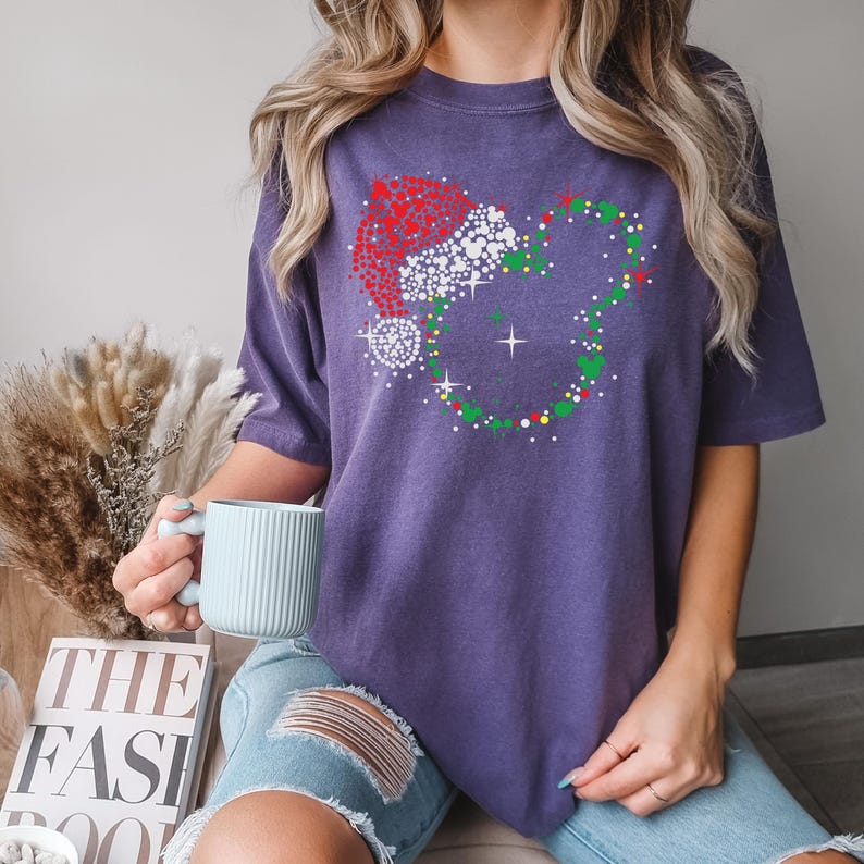 Puede incluir: Camiseta morada con un dise&ntilde;o tem&aacute;tico navide&ntilde;o. El dise&ntilde;o incluye un gorro de Pap&aacute; Noel y una corona hecha de puntos y brillos. La camiseta se combina con vaqueros azules y una taza azul claro.
