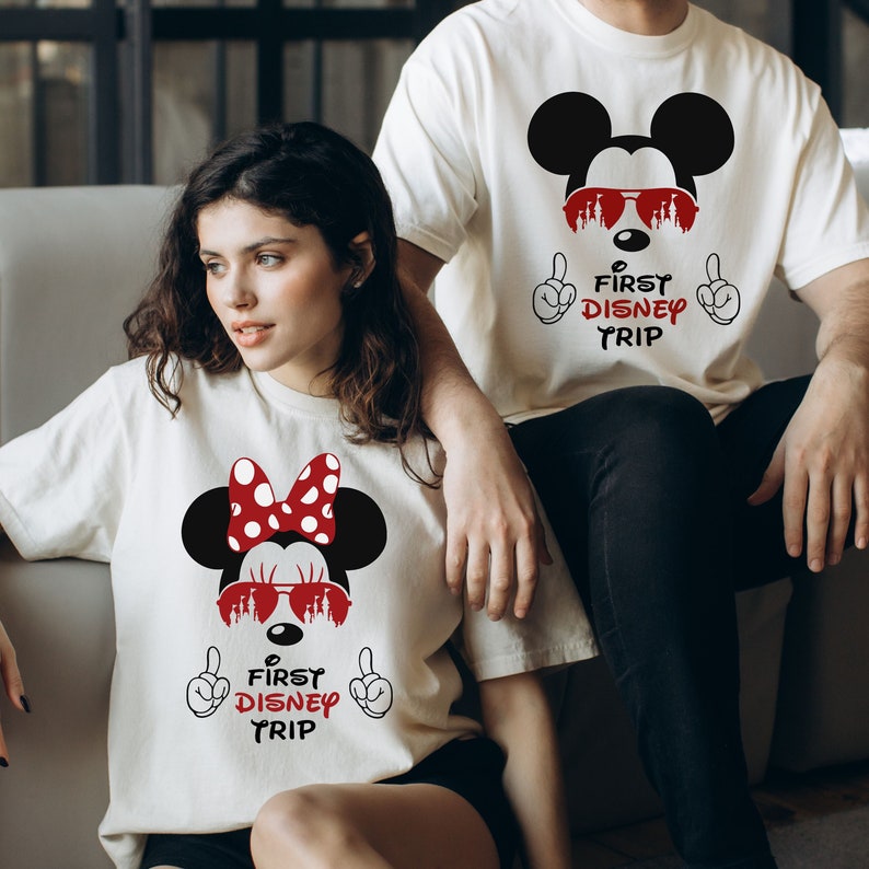 May include: White t-shirts with a Mickey Mouse and Minnie Mouse graphic. The graphic features Mickey Mouse wearing sunglasses and Minnie Mouse wearing a red bow and sunglasses. The text on the shirts reads "First Disney Trip".
