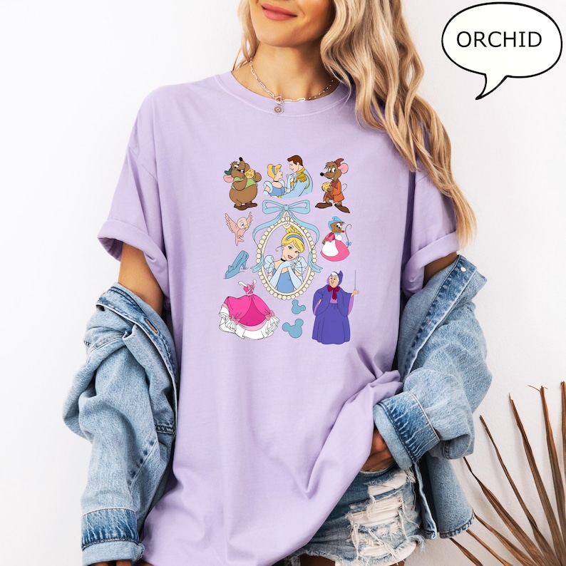 Puede incluir: Una camiseta de color lila claro con un dise&ntilde;o de dibujos animados con personajes de Cenicienta. El dise&ntilde;o incluye a Cenicienta, ratones, p&aacute;jaros y otros personajes. La palabra "ORCHID" est&aacute; en un bocadillo.