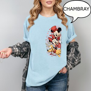 Puede incluir: Camiseta azul claro con un gr&aacute;fico de Mickey Mouse, Minnie Mouse y Pluto con atuendos de San Valent&iacute;n. Mickey lleva un sombrero de copa, Minnie un lazo y Pluto un collar de reno. La palabra "CHAMBRAY" est&aacute; en una burbuja.
