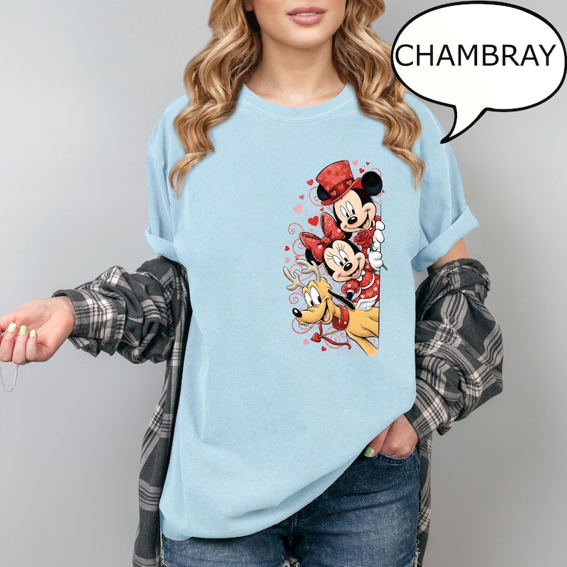 Puede incluir: Camiseta azul claro con un gr&aacute;fico de Mickey Mouse, Minnie Mouse y Pluto con atuendos de San Valent&iacute;n. Mickey lleva un sombrero de copa, Minnie un lazo y Pluto un collar de reno. La palabra "CHAMBRAY" est&aacute; en una burbuja.