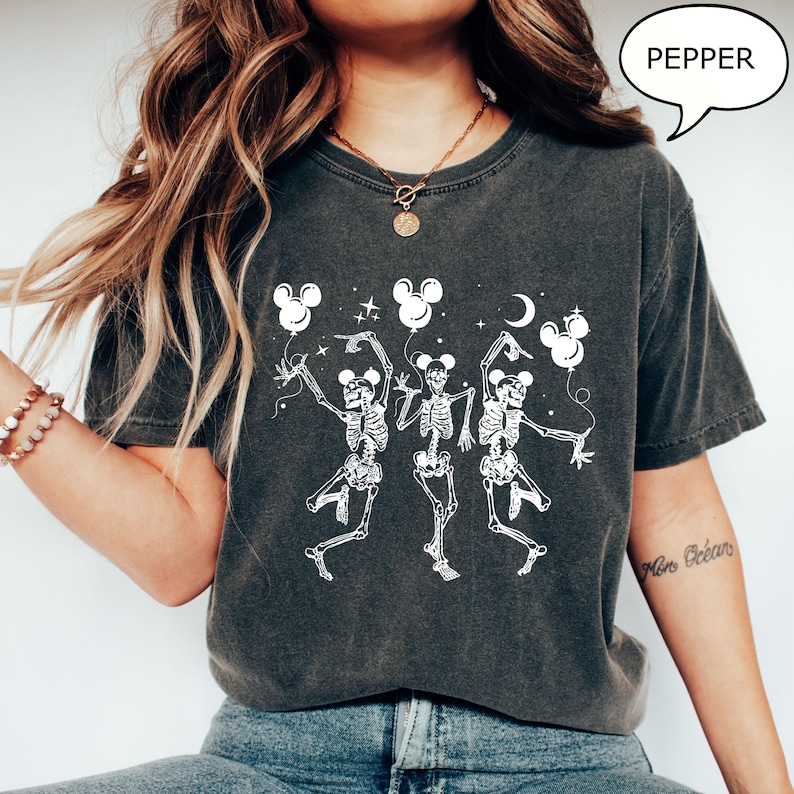 May include: Dark gray t-shirt featuring a white graphic of three dancing skeletons holding Mickey Mouse-shaped balloons. The word "PEPPER" is in a speech bubble. The design includes stars and a crescent moon.