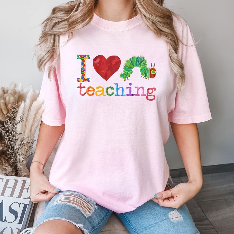 Op de afbeelding: Een roze T-shirt met de tekst "Ik hou van lesgeven" en een kleurrijke illustratie van een rups met een hart.