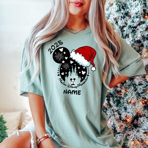 Peut inclure: Un t-shirt vert clair avec une silhouette noire et blanche de Mickey Mouse portant un chapeau de P&egrave;re No&euml;l. La silhouette comprend des feux d'artifice, le ch&acirc;teau de Cendrillon et le texte "Making Disney Memories 2025". Le texte "NAME" est sous la silhouette.