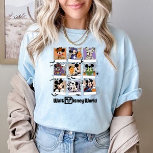 以下が含まれることがあります： 薄い青色のTシャツに、ミッキーマウスがさまざまなハロウィーンのコスチュームを着ているグラフィックがプリントされています。グラフィックは9つの正方形のグリッドに配置されており、それぞれ異なるコスチュームが描かれています。グラフィックの下には「Walt Disney World」というテキストが書かれています。