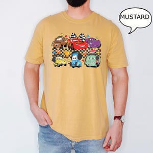 May include: A mustard yellow t-shirt with a cartoon graphic of a group of cars from the Disney Pixar movie Cars. The cars are on a checkered flag background with stars.