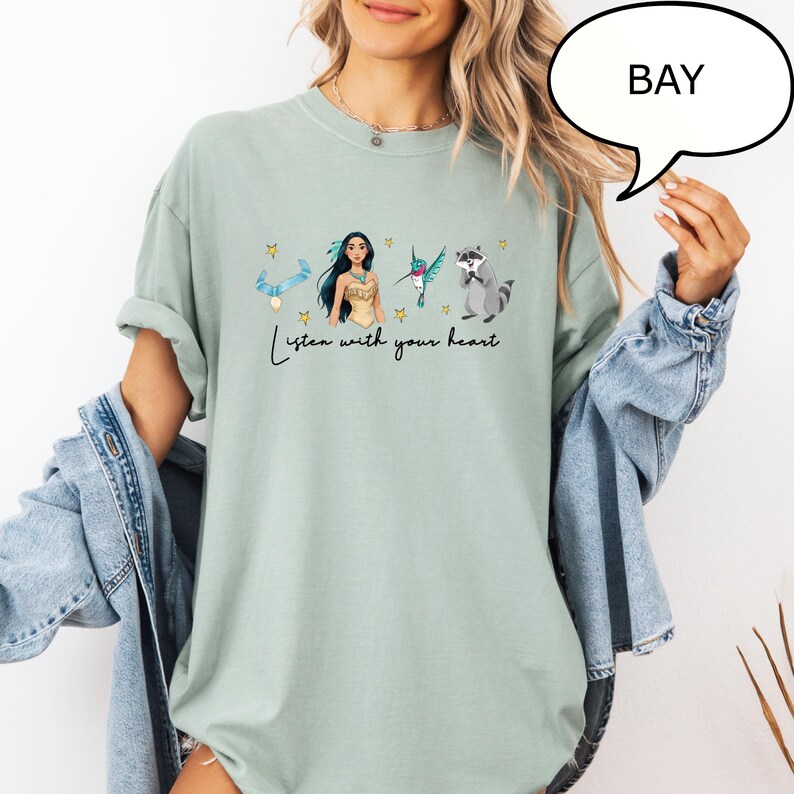K&ouml;nnte beinhalten: Hellgr&uuml;nes T-Shirt mit einer Grafik einer amerikanischen Ureinwohnerin, eines Kolibris und eines Waschb&auml;ren, mit dem Text "Listen with your heart". Das Design umfasst Sterne und ein blaues Band. Das T-Shirt hat eine lockere Passform.