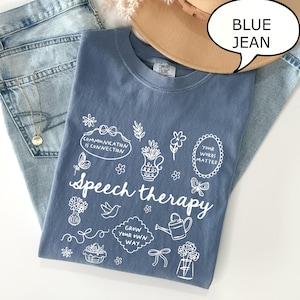 May include: A blue Comfort Colors t-shirt with the words "Speech therapy" and various white illustrations. The shirt is paired with blue jeans and a tan hat. The text "Blue Jean" is in a speech bubble.