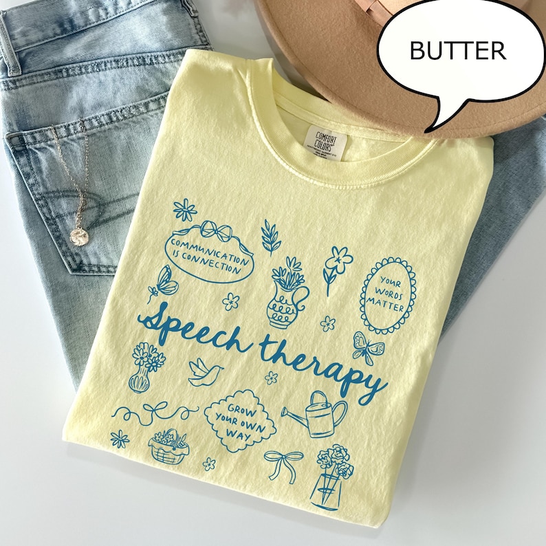 May include: A light yellow t-shirt with the words "Speech therapy" and various illustrations in blue. The shirt also features the phrases "Communication is connection", "Your words matter", and "Grow your own way". The shirt is laid on top of blue jeans.