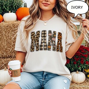 May include: Ivory-colored t-shirt with the word "MAMA" in a bold, decorative font. The letters feature leopard print, plaid, and floral patterns with pumpkins. The shirt is paired with blue jeans.