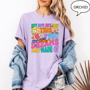 Może przedstawiać: Fioletowy t-shirt z nadrukiem graficznym przedstawiającym dwie kresk&oacute;wkowe dziewczyny i tekst "Hey Now Hey Now This Is What Dreams Are Made Of".
