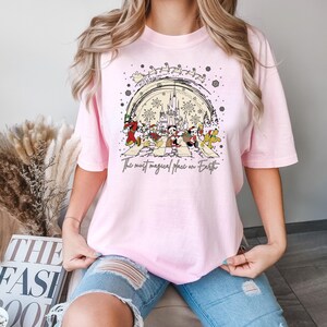 Puede incluir: Camiseta rosa con un gr&aacute;fico de Mickey Mouse y sus amigos caminando por una calle con el texto "The most magical place on Earth" y un dibujo del castillo de Cenicienta en el fondo.