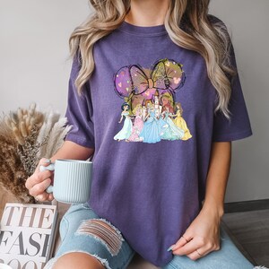 Puede incluir: Camiseta morada con un dise&ntilde;o caprichoso de un lazo con orejas de rat&oacute;n y varias princesas. El lazo est&aacute; decorado con corazones y siluetas de Mickey Mouse. Las princesas visten vestidos coloridos.